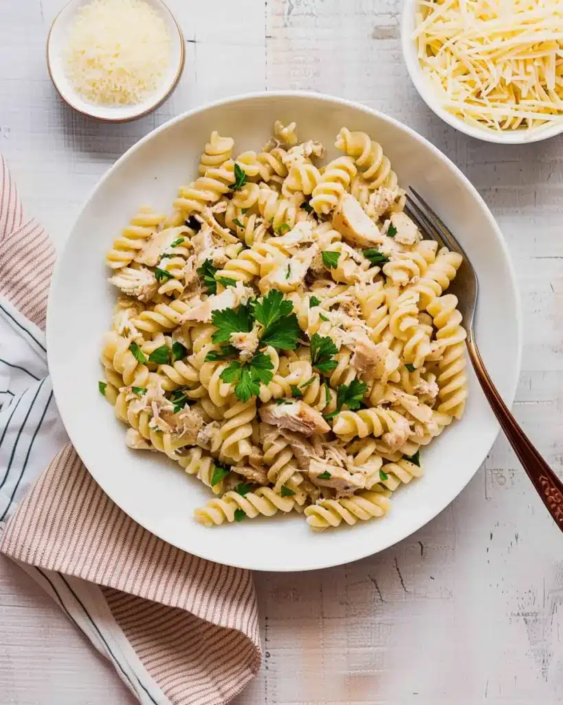 Creamy garlic parmesan chicken pasta with rotini noodles, served in a white bowl and topped with parsley, on a rustic wooden table