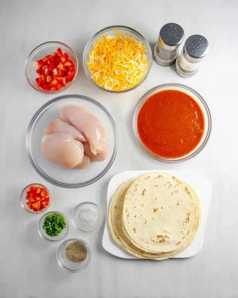 Overhead view of chicken enchilada casserole ingredients including raw chicken, cheese, tortillas, enchilada sauce, diced tomatoes, and spices on a gray kitchen background.