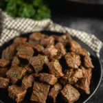 Slow-cooked steak bites on a plate with herb garnish and rustic kitchen background
