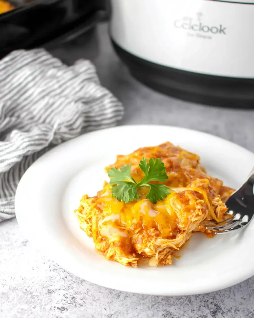 A serving of slow cooker chicken enchilada casserole topped with melted cheese on a white plate, with a Crockpot in the background.