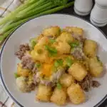 Overhead view of slow cooker cowboy casserole served with crispy tater tots, ground beef, melted cheese, and green onions on a white plate
