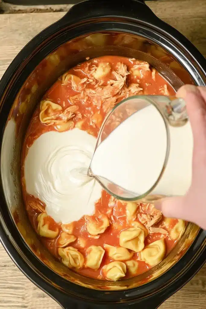 Pouring heavy cream into a slow cooker filled with chicken, tortellini, and tomato sauce