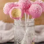 Bright pink cake pops with white sprinkles in a glass jar, one cake pop with a bite showing moist vanilla cake inside.