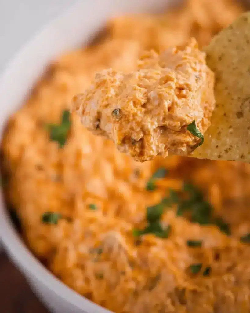 Close-up of tortilla chip scooping creamy buffalo chicken dip with herbs in background