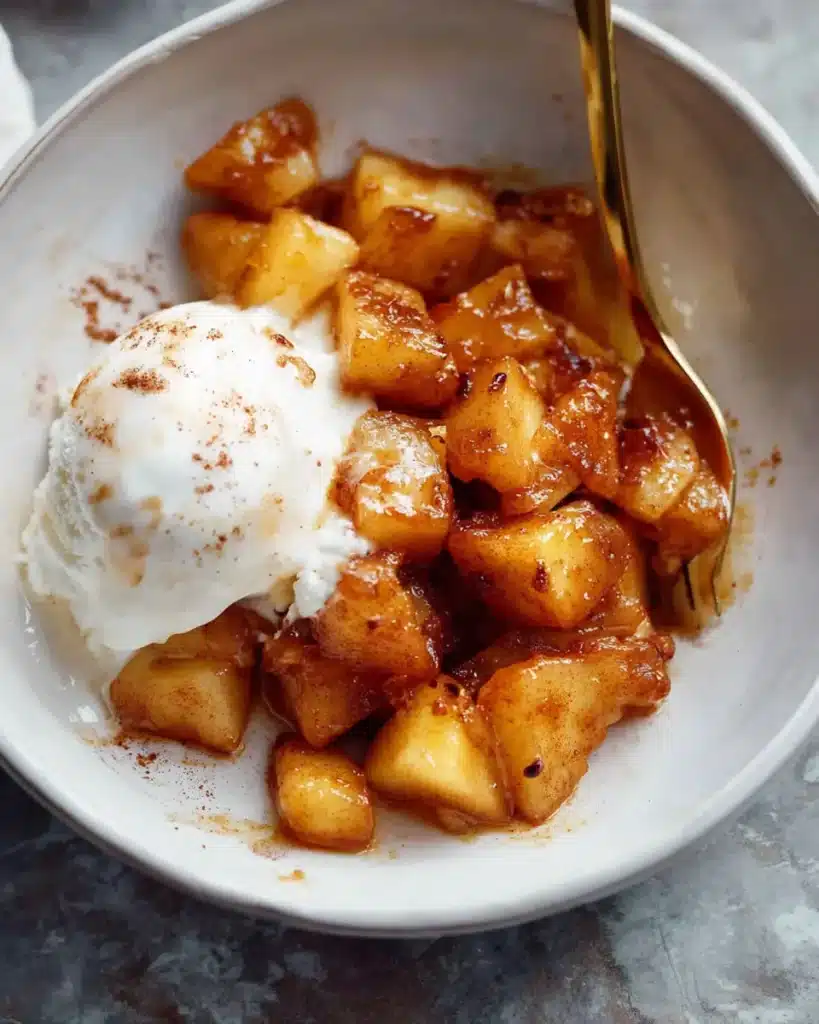 Caramelized cinnamon apple chunks with melting vanilla ice cream and gold spoon in white bowl