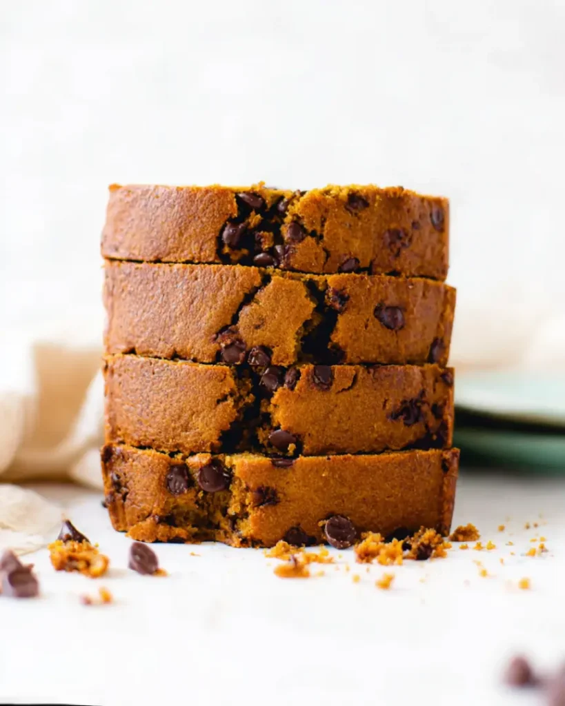 Stack of moist chocolate chip pumpkin bread slices with melty chocolate chips on a ceramic surface.