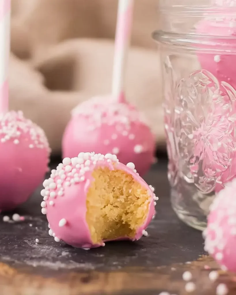 Bright pink cake pops with white sprinkles resting on a slate surface, one bitten open showing moist vanilla cake inside.