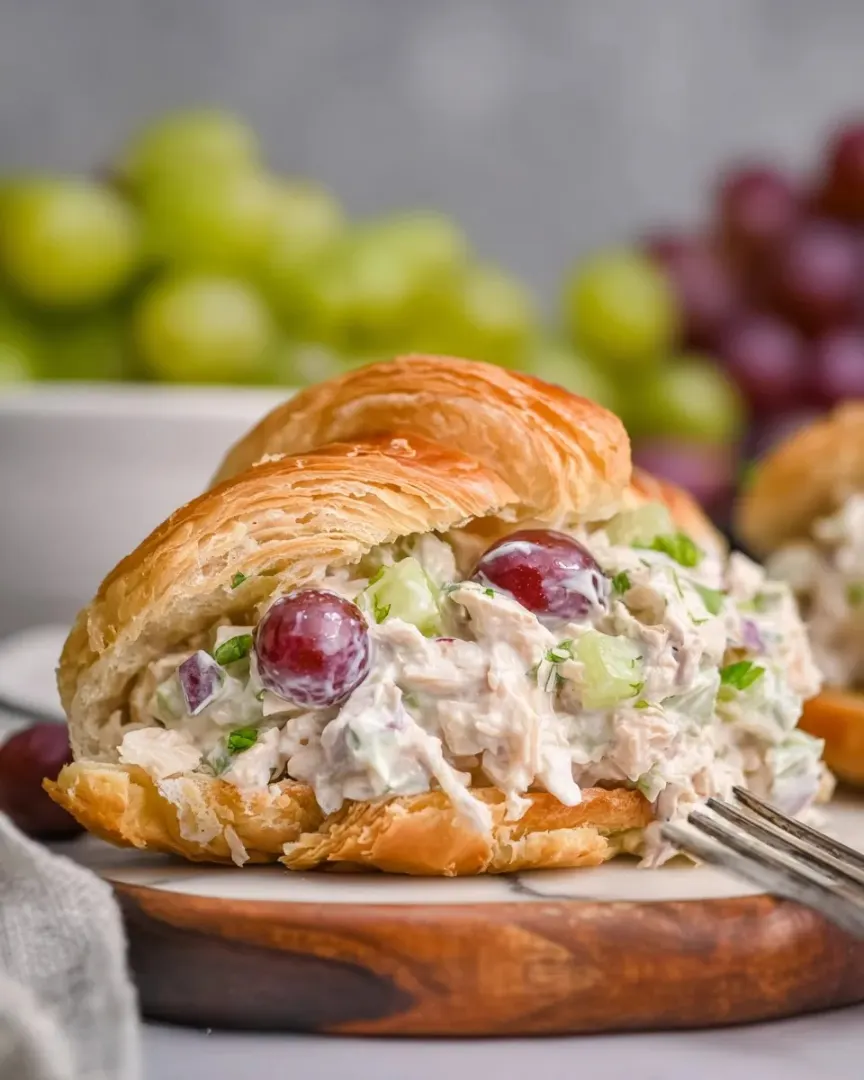 Chicken salad croissant sandwich filled with grapes, celery, and red onion on a wooden board