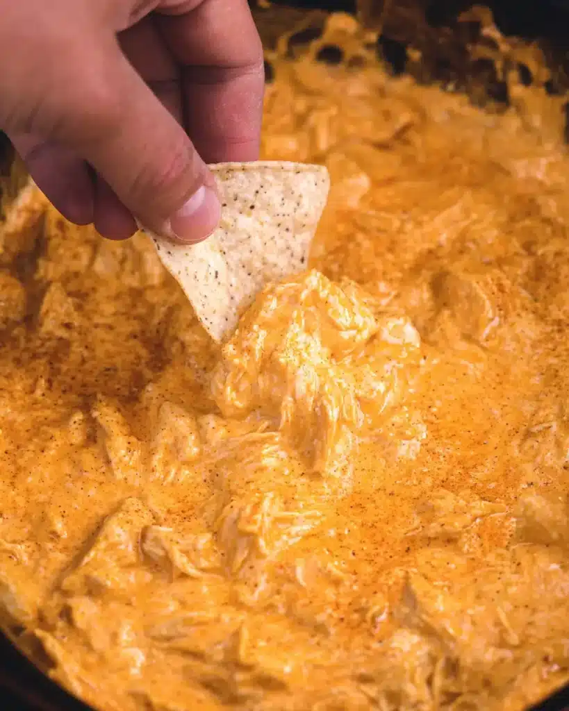 Hand dipping a tortilla chip into creamy buffalo chicken dip in a slow cooker with rich orange cheesy texture.