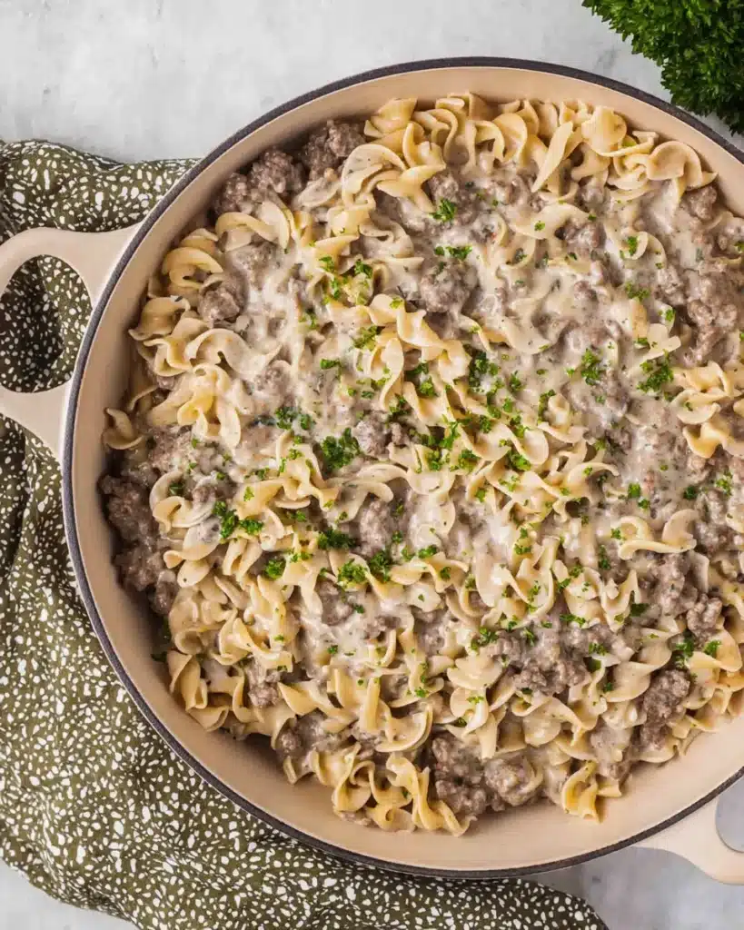 Creamy ground beef stroganoff in a skillet with egg noodles and fresh parsley, viewed from above on a light surface.