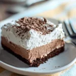 Rich layered French Silk Brownie with whipped cream and chocolate shavings on a ceramic plate.