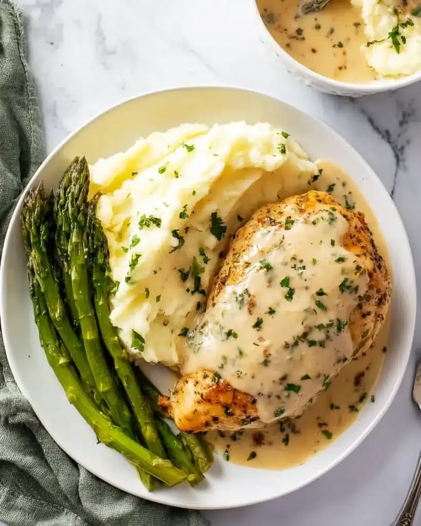 Garlic Parmesan Chicken served with mashed potatoes and roasted asparagus, all topped with creamy parmesan sauce on a white plate.