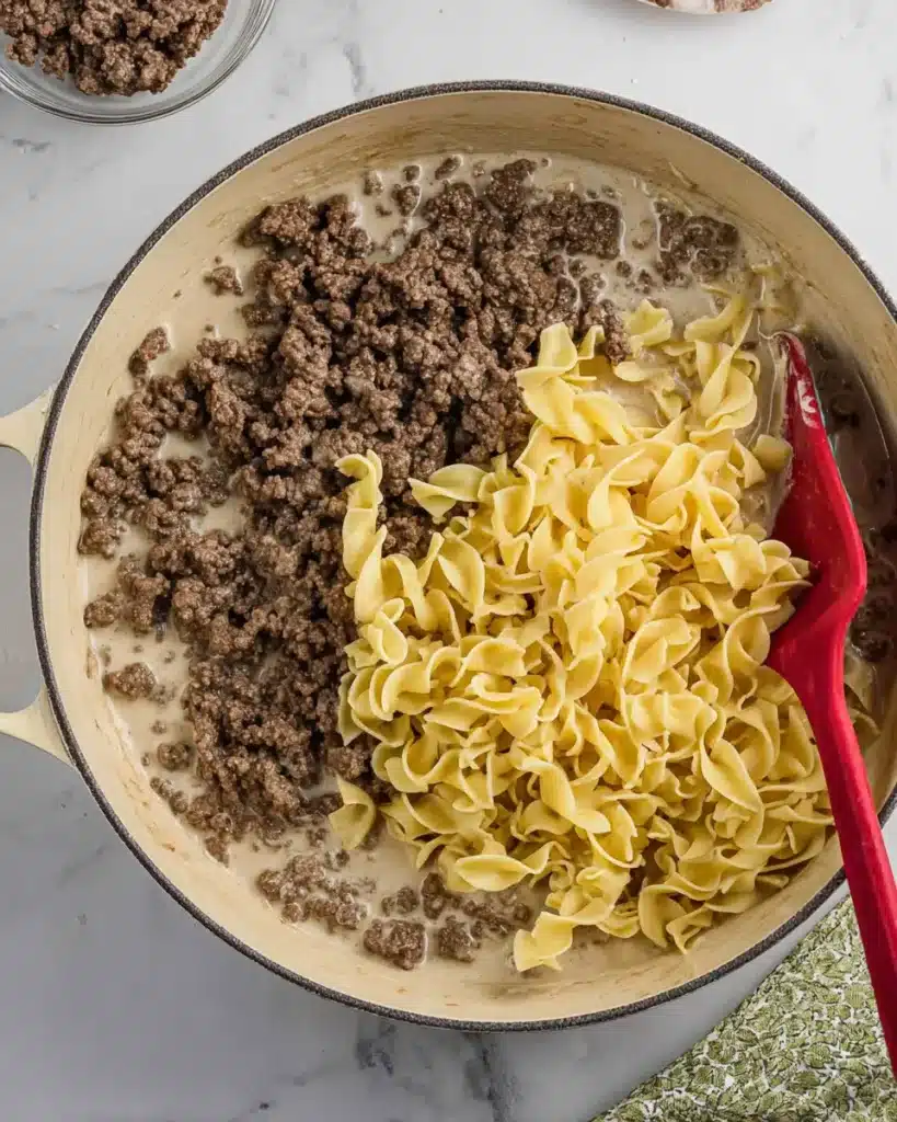 Ground beef and egg noodles added to creamy sauce in a skillet, ready to be stirred for stroganoff