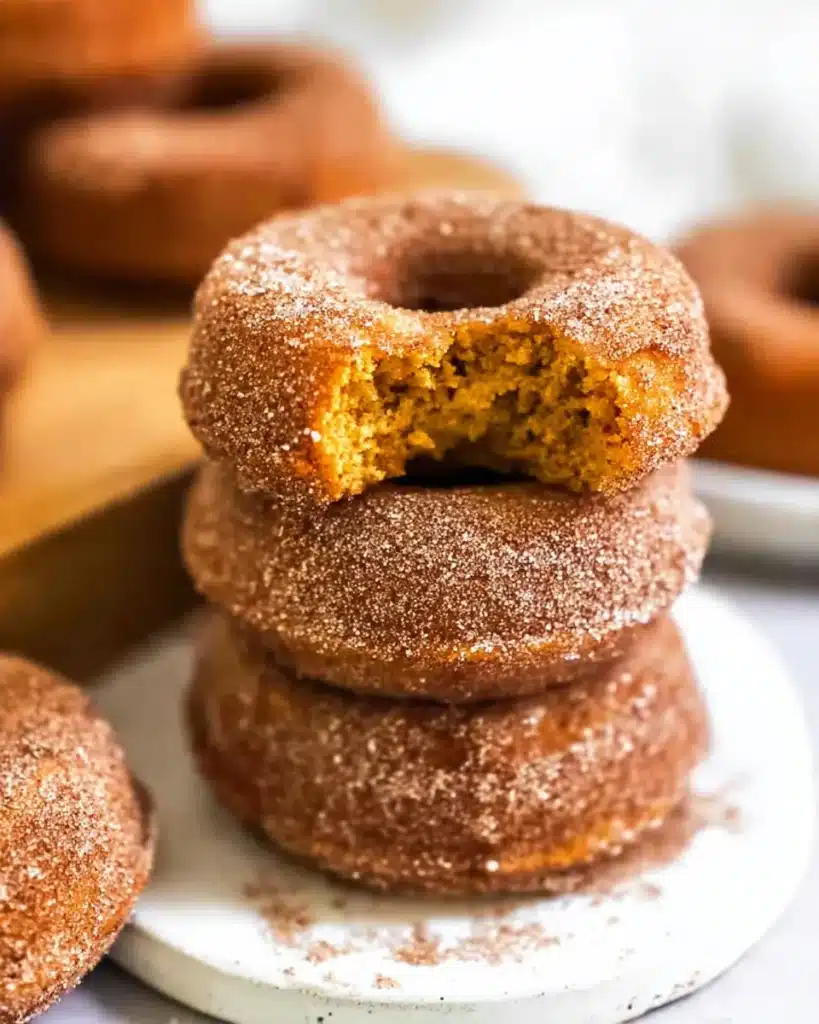 Stack of baked pumpkin donuts coated in cinnamon sugar with a bite showing soft pumpkin interior