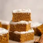Stack of fluffy pumpkin bars with cream cheese frosting on a rustic table with cinnamon sticks