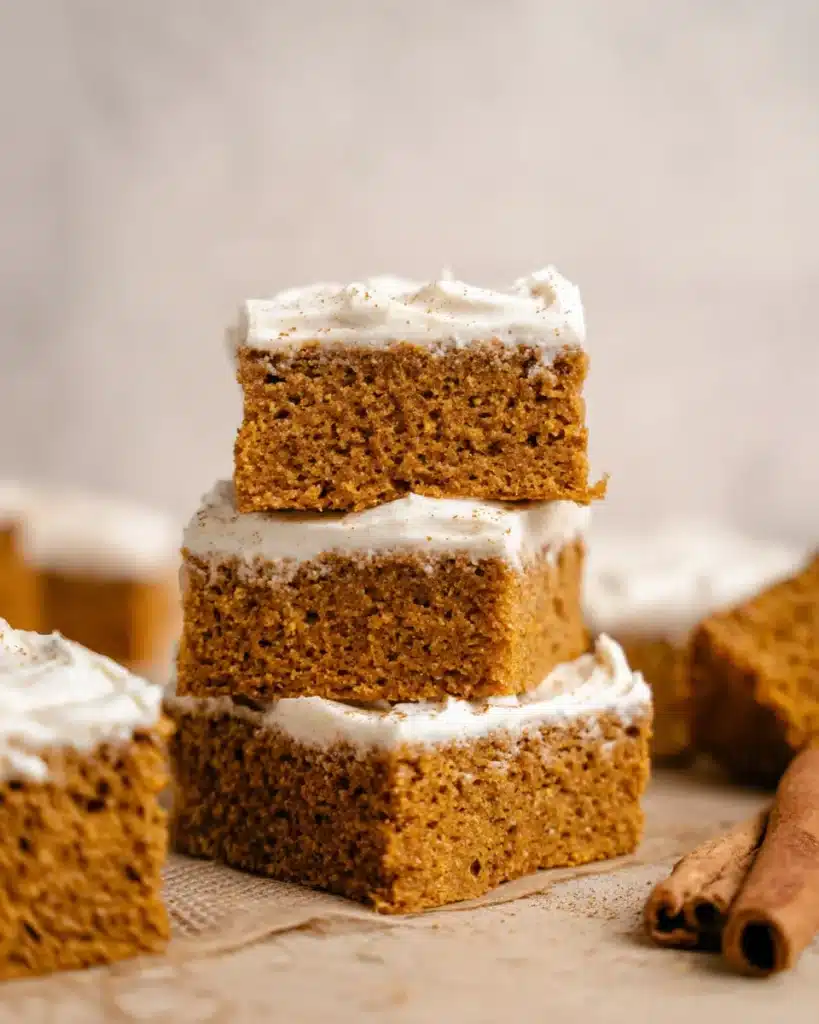 Stack of fluffy pumpkin bars with cream cheese frosting on a rustic table with cinnamon sticks