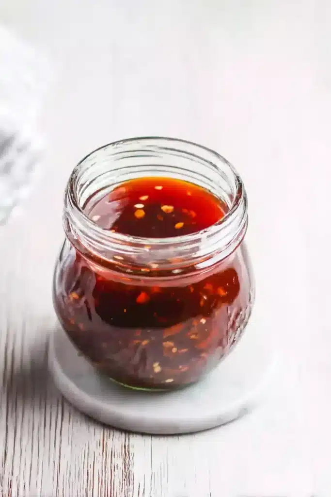 Jar of homemade sticky sesame garlic sauce with garlic and chili bits in a glass jar on a whitewashed wooden table.
