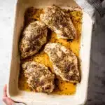 Juicy baked chicken breasts in a white ceramic baking dish with golden pan juices and herb seasoning.