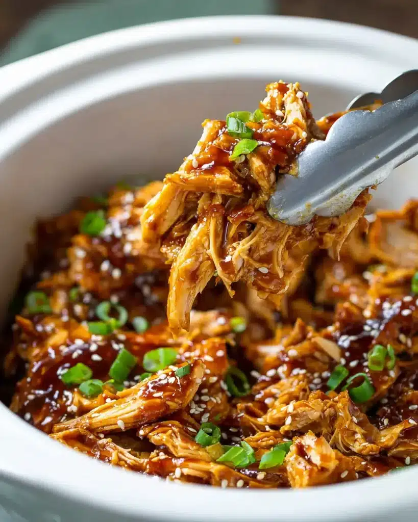 Tongs lifting glazed shredded teriyaki chicken from white slow cooker with sesame and green onion