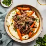 Overhead view of slow cooker garlic herb pot roast with carrots and gravy over mashed potatoes, garnished with fresh parsley