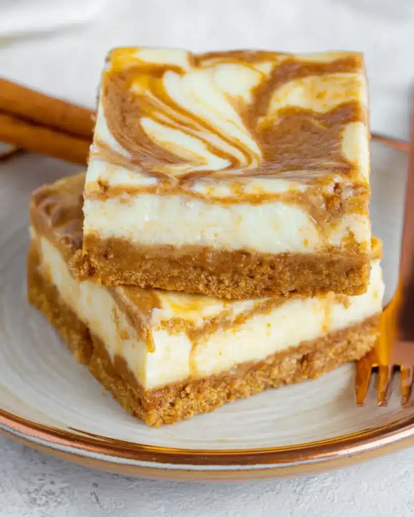Pumpkin cheesecake bars with a creamy swirl on a crumb crust, stacked on a plate with a gold fork and cinnamon stick