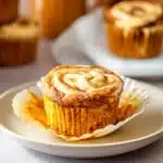 Fresh pumpkin cream cheese swirl muffin with creamy topping on white plate, unwrapped and ready to eat