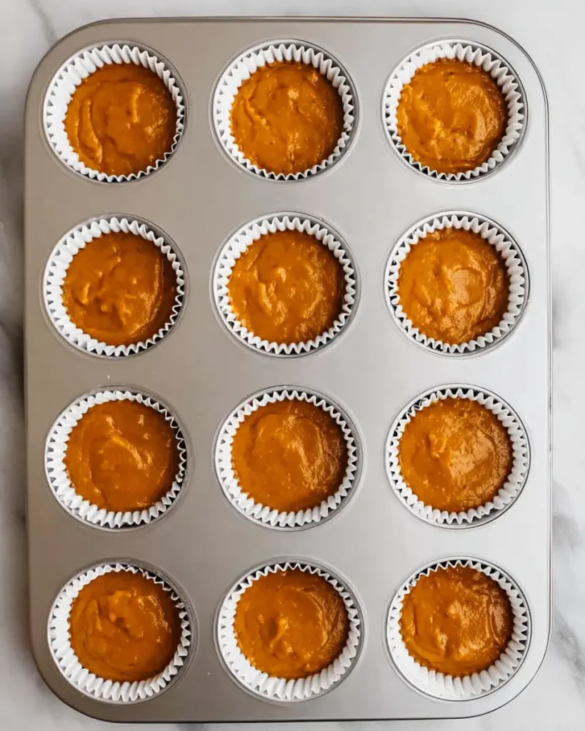 Muffin tin filled with pumpkin cupcake batter in white liners on marble surface