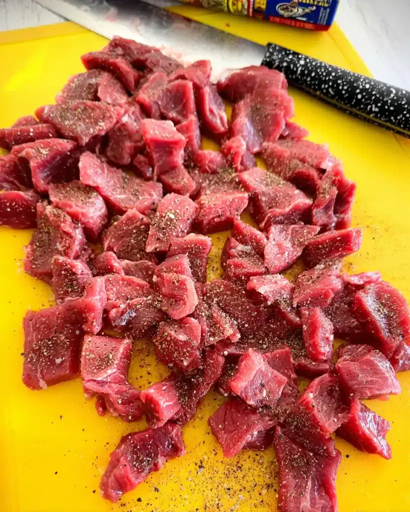 Close-up of raw steak strips on yellow cutting board, seasoned with coarse steak seasoning, knife nearby, ready for cooking.
