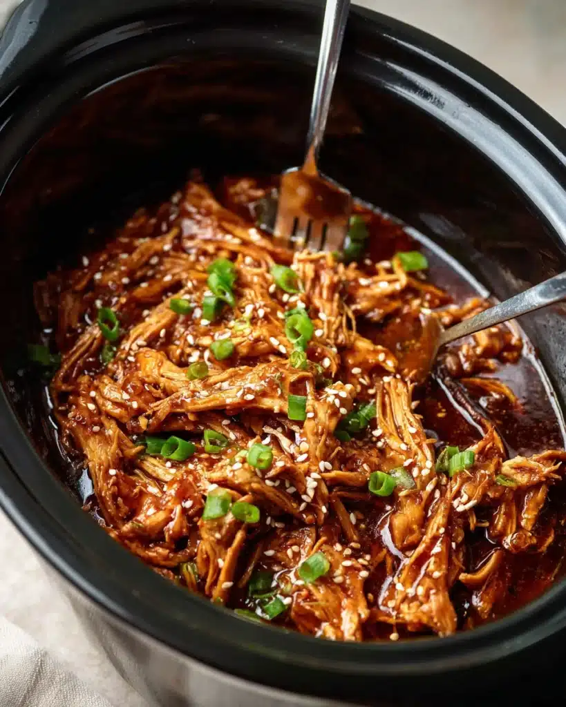 Slow cooker filled with shredded honey garlic chicken, topped with sesame seeds and green onions, being pulled apart with forks.