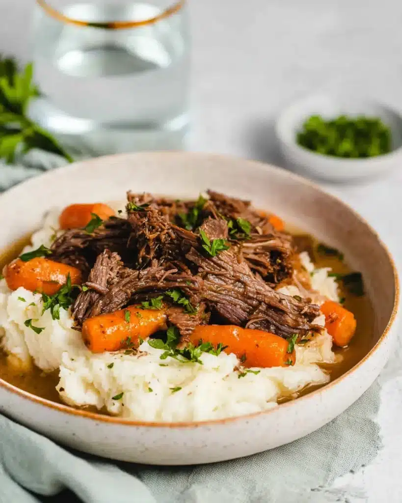 Side view of a bowl of slow cooker garlic herb pot roast with baby carrots and mashed potatoes, garnished with parsley