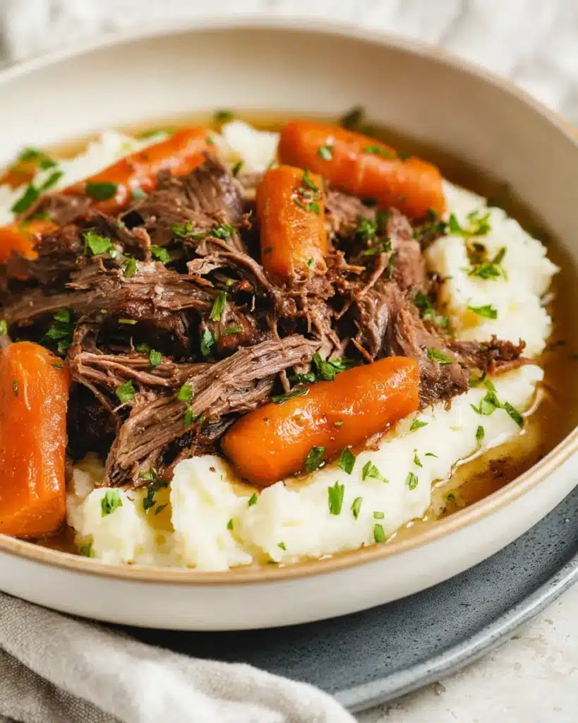 Shredded garlic herb pot roast served over creamy mashed potatoes with baby carrots and fresh parsley in a rustic bowl.
