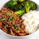A bowl of slow cooker honey garlic chicken topped with sesame seeds and green onions, served with white rice and roasted broccoli.