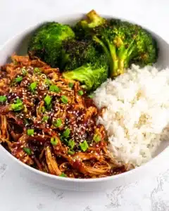 A bowl of slow cooker honey garlic chicken topped with sesame seeds and green onions, served with white rice and roasted broccoli.