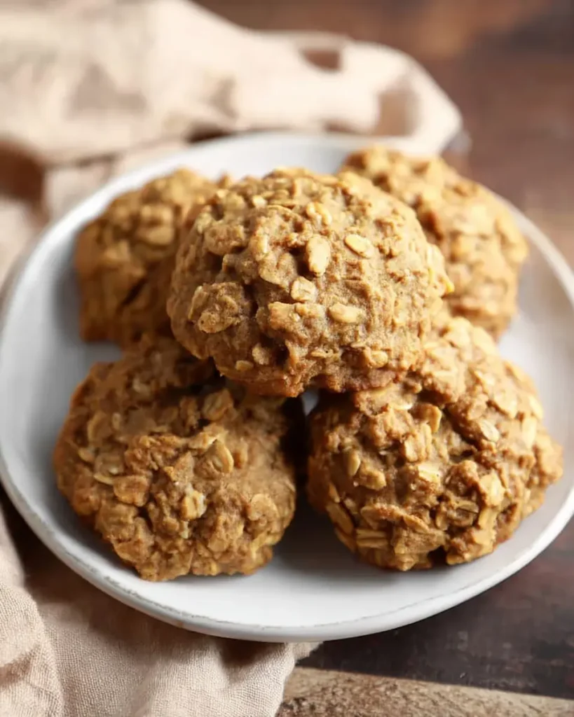 Five soft pumpkin oatmeal cookies with visible oats and a chunky cracked texture, served on a white ceramic plate over a beige linen napkin and dark wood table.