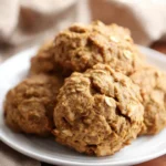 Soft pumpkin oatmeal cookies stacked on a white plate, showing a golden-brown, chewy texture with visible oats and cracks, set on a rustic wooden table with a brown fabric napkin in soft natural light.