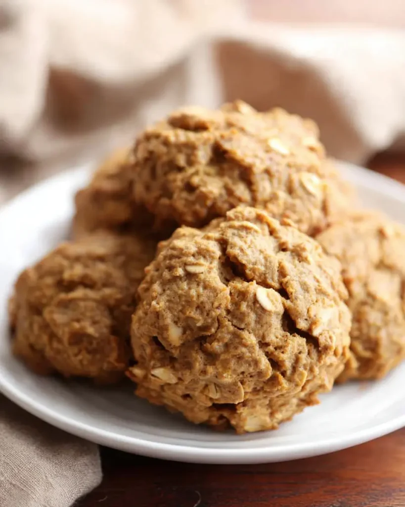 Soft pumpkin oatmeal cookies stacked on a white plate, showing a golden-brown, chewy texture with visible oats and cracks, set on a rustic wooden table with a brown fabric napkin in soft natural light.