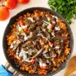 Overhead view of Steak and Queso Rice skillet with juicy steak strips, fluffy tomato rice, queso drizzle, and fresh parsley, surrounded by tomatoes and rustic table setting.