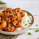 Bowl of sticky sesame cauliflower with white rice, garnished with green onions and sesame seeds on a rustic wooden table.