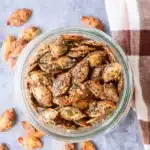 Crunchy roasted pumpkin seeds in a glass jar, coated with ranch seasoning and herbs, on a cozy striped cloth