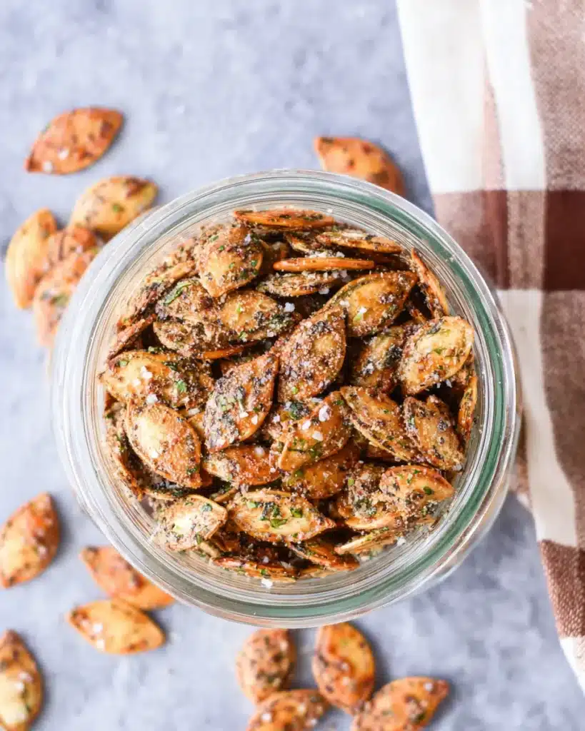 Crunchy roasted pumpkin seeds in a glass jar, coated with ranch seasoning and herbs, on a cozy striped cloth