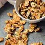 Air Fryer Pumpkin Seeds spilled from white bowl with herb seasoning on beige background”