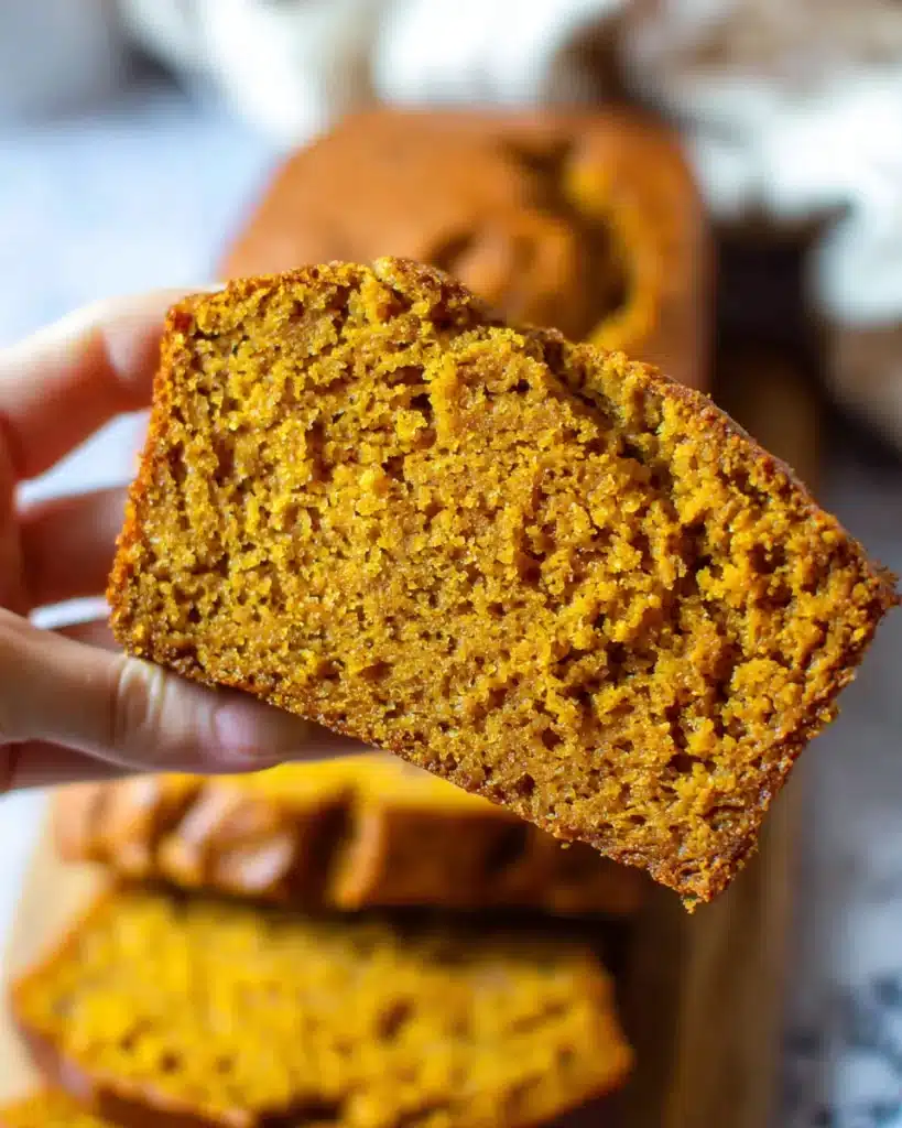 Hand holding a slice of moist vanilla pudding pumpkin bread showing tender orange crumb and golden crust