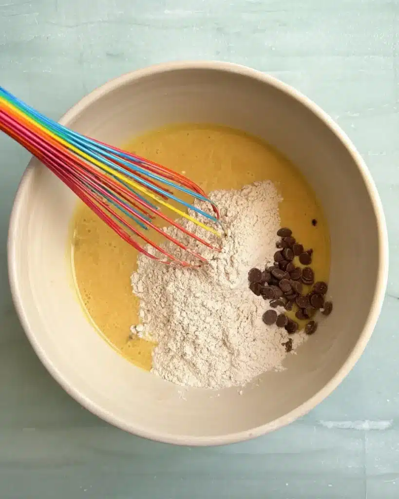 Bowl with batter, flour, chocolate chips, and a colorful rainbow whisk.