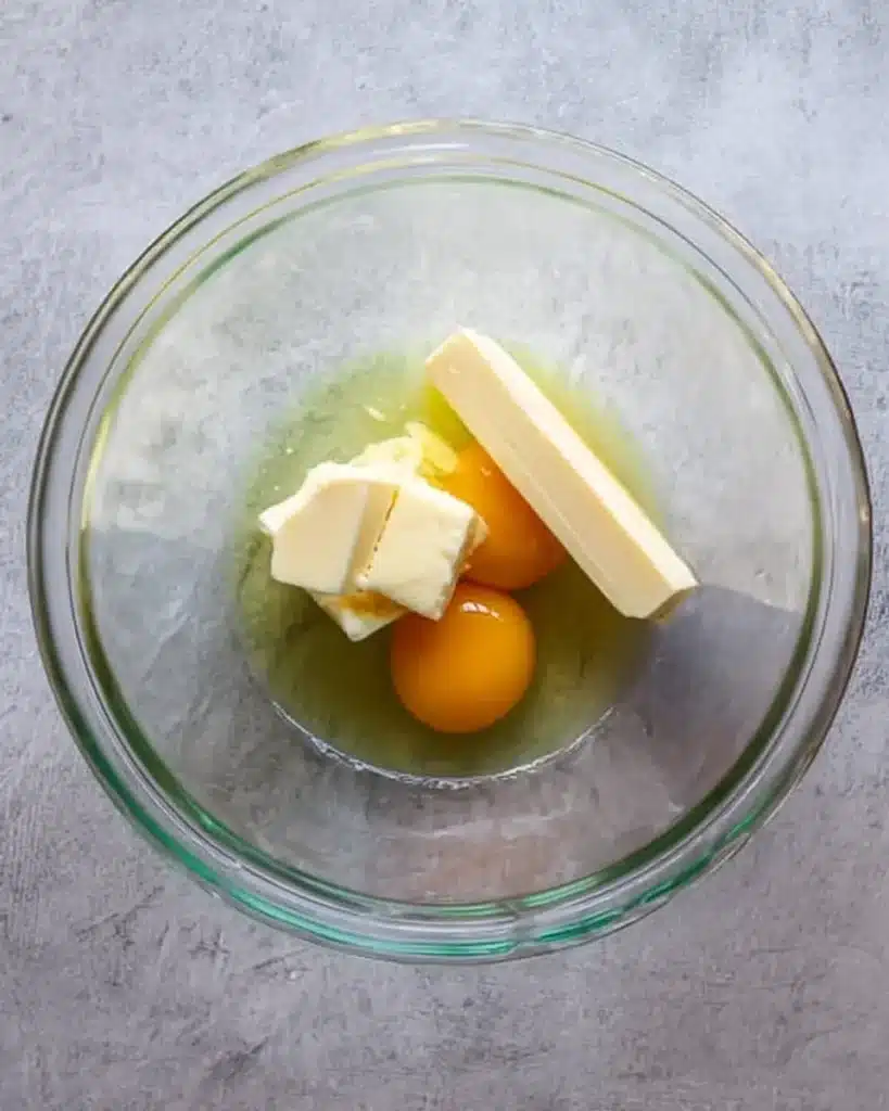 Two whole eggs and butter sticks in a glass bowl on a light gray kitchen surface