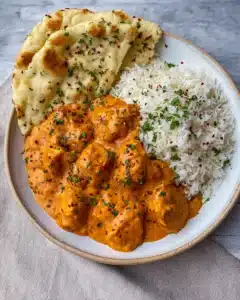 Overhead view of creamy butter chicken with naan and rice on a ceramic plate with herb garnish