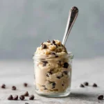 Glass jar of edible cookie dough with chocolate chips and spoon on a stone surface