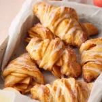 Close-up of golden crescent roll apple turnovers with vanilla glaze on parchment paper and sliced apples in a white tray
