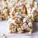 Close-up of gooey funfetti marshmallow popcorn treats with rainbow sprinkles and white chocolate chips on a light surface