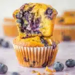 Blueberry cottage cheese muffins stacked with one torn open to show a fluffy golden interior and juicy blueberries, surrounded by fresh berries on a white surface