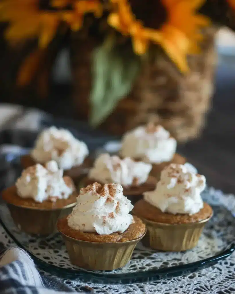 Easy pumpkin pie cupcakes topped with whipped cream, arranged on a glass tray with cozy fall decor and rustic lace doily